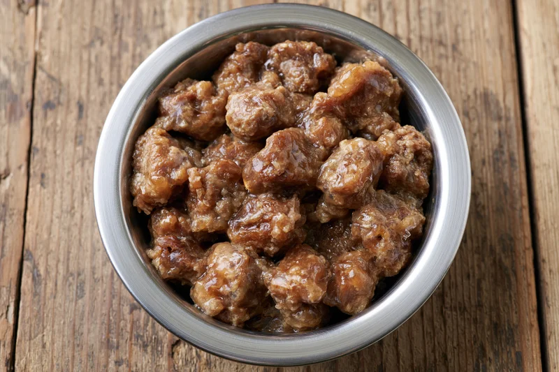 Wet chunky dog food in a stainless steel bowl on a wooden surface