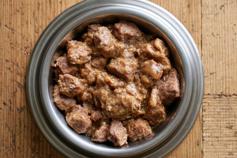 Wet chunky dog food in a stainless steel bowl on wooden surface