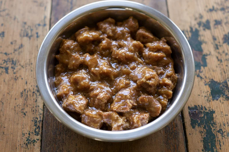 Wet chunky dog food in a stainless steel bowl on a wooden table