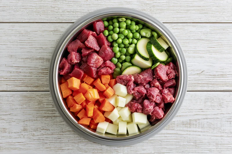 Raw dog food ingredients arranged in a stainless steel bowl on a white wooden surface