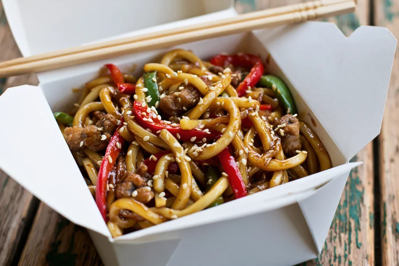 Stir-fried udon noodles with pork and bell peppers in a takeout box on a wooden table