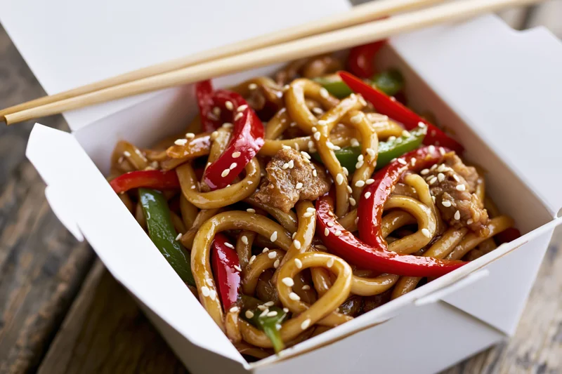 Stir-fried udon noodles with pork and bell peppers in a takeout box on a wooden surface