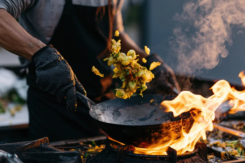 Cook tossing vegetables in a wok over an open flame outdoors