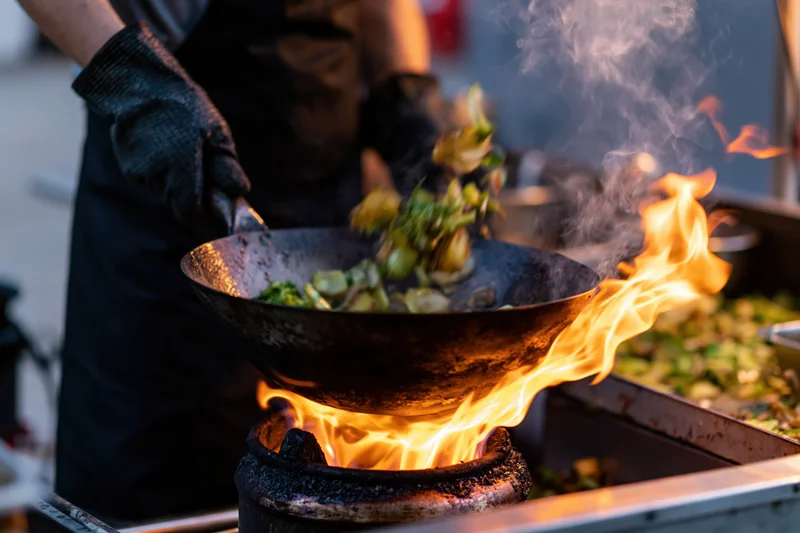 Chef tossing vegetables in a wok over an open flame at a street food stall