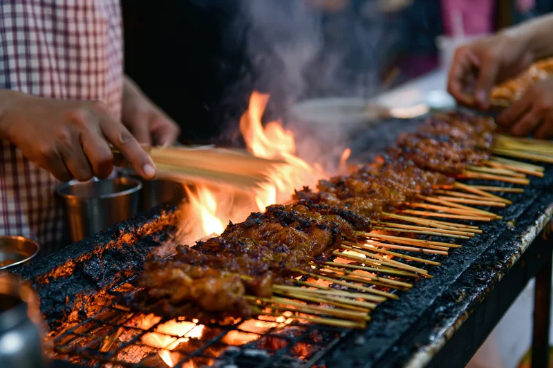 Vendor grilling rows of meat satay skewers over open flame at street market