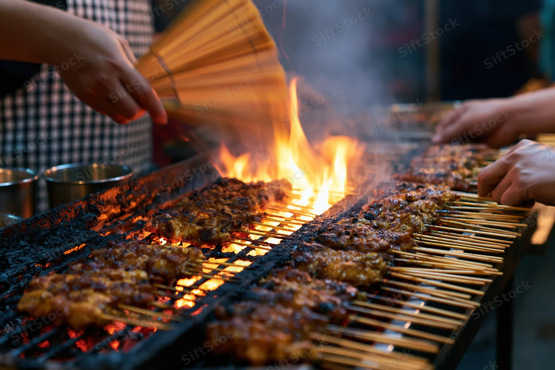 Hands grilling satay skewers over open flame at night market stall
