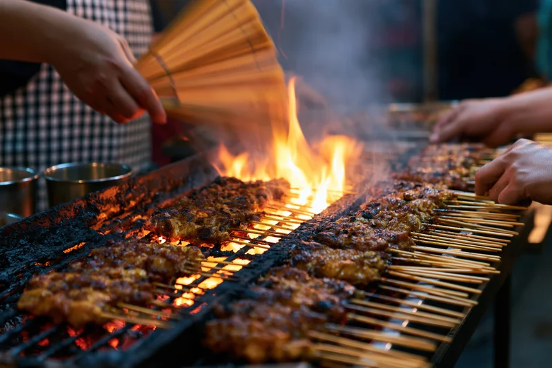 Hands grilling satay skewers over open flame at night market stall