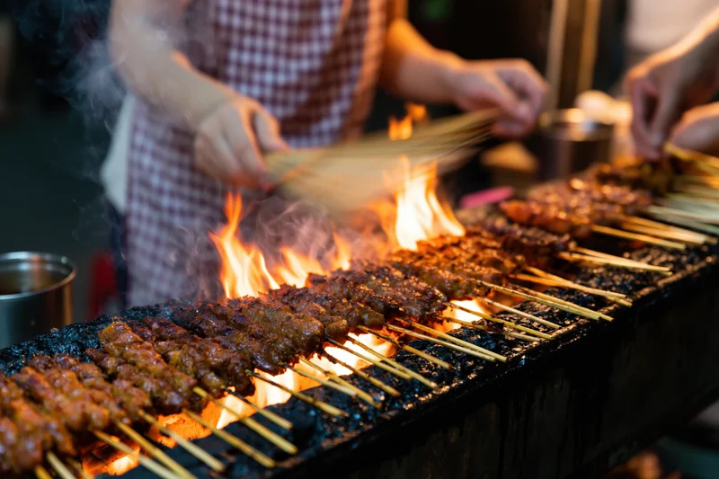 Vendor grilling rows of meat satay skewers over open flame at night market