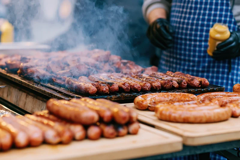 Sausages grilling on a barbecue with smoke rising as a vendor in apron and gloves prepares them outdoors