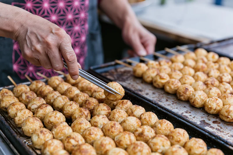Vendor hands flipping rows of takoyaki on a grill at a street food market