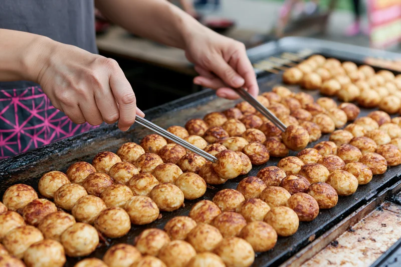Hands flipping rows of takoyaki on a griddle at a street food market