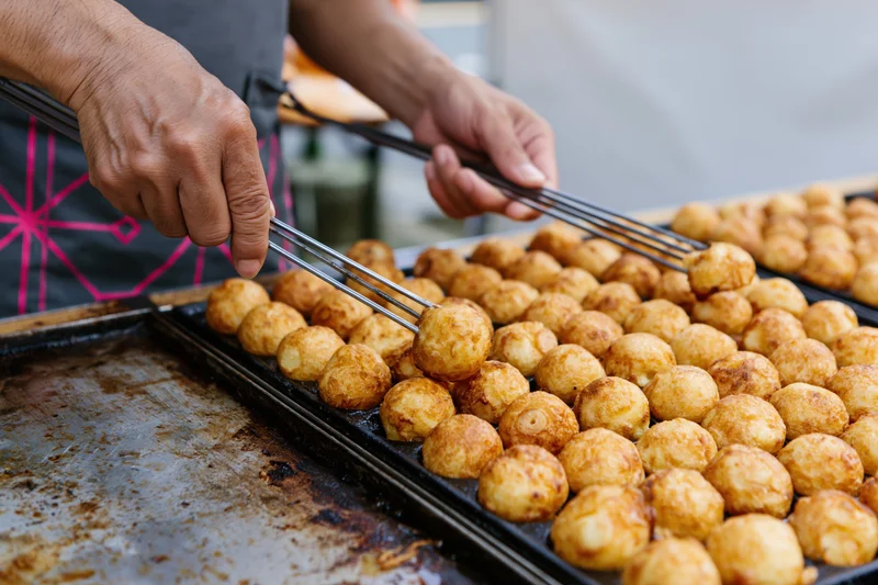 Vendor hands flipping rows of takoyaki on a griddle at a street food stall