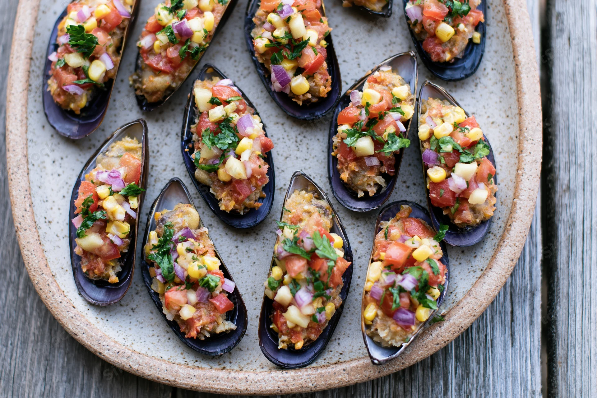 Stuffed mussels on half shells topped with chopped tomato and corn on a ceramic plate on wooden table