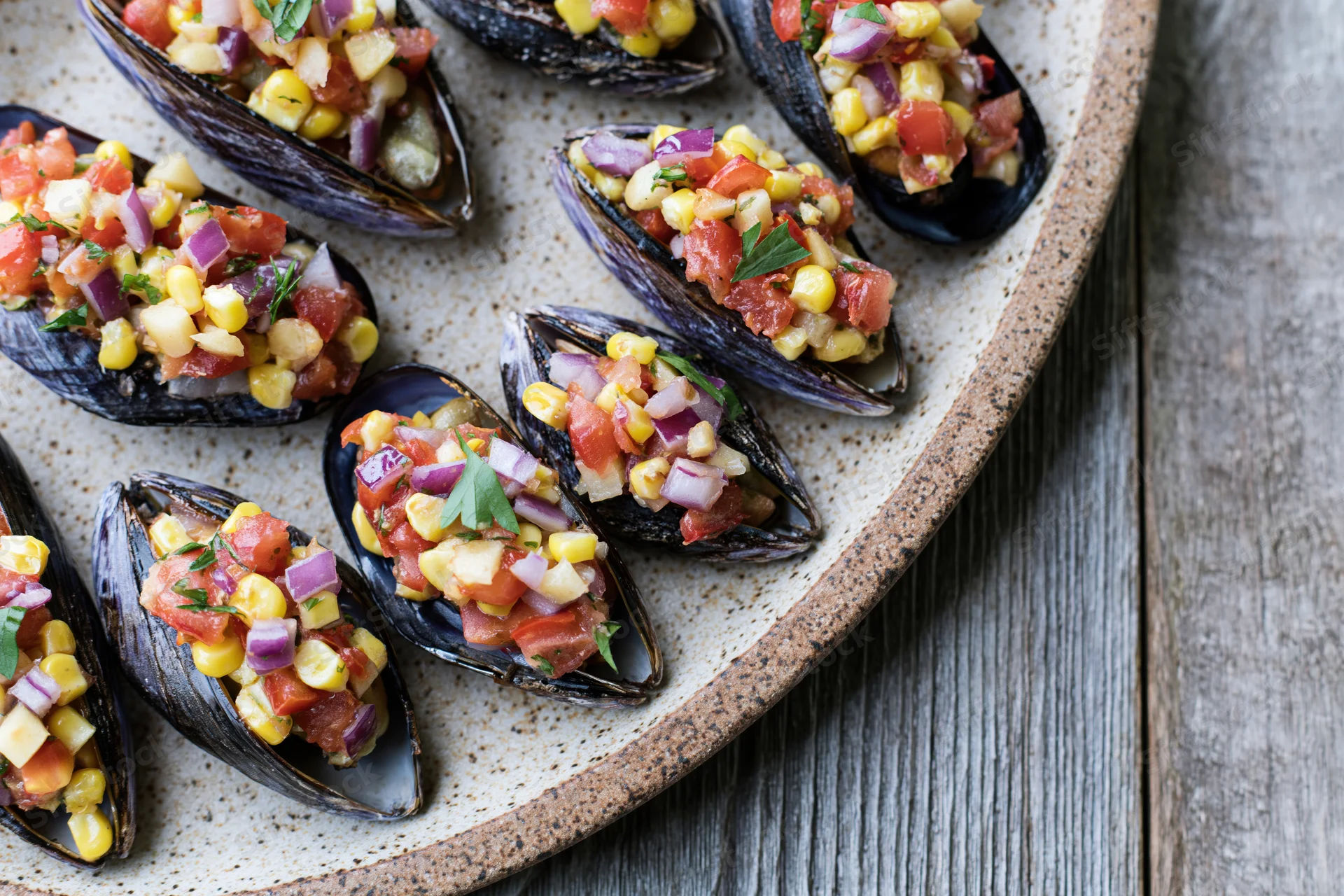 Stuffed mussels on half shells topped with tomato and corn salsa served on a speckled ceramic plate on a wooden table