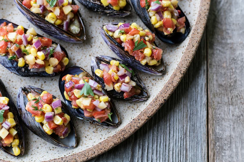 Stuffed mussels on half shells topped with tomato and corn salsa served on a speckled ceramic plate on a wooden table