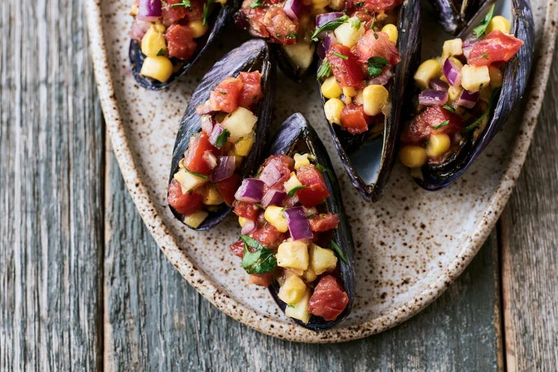 Stuffed mussels on half shells topped with tomato and corn salsa on a speckled ceramic plate