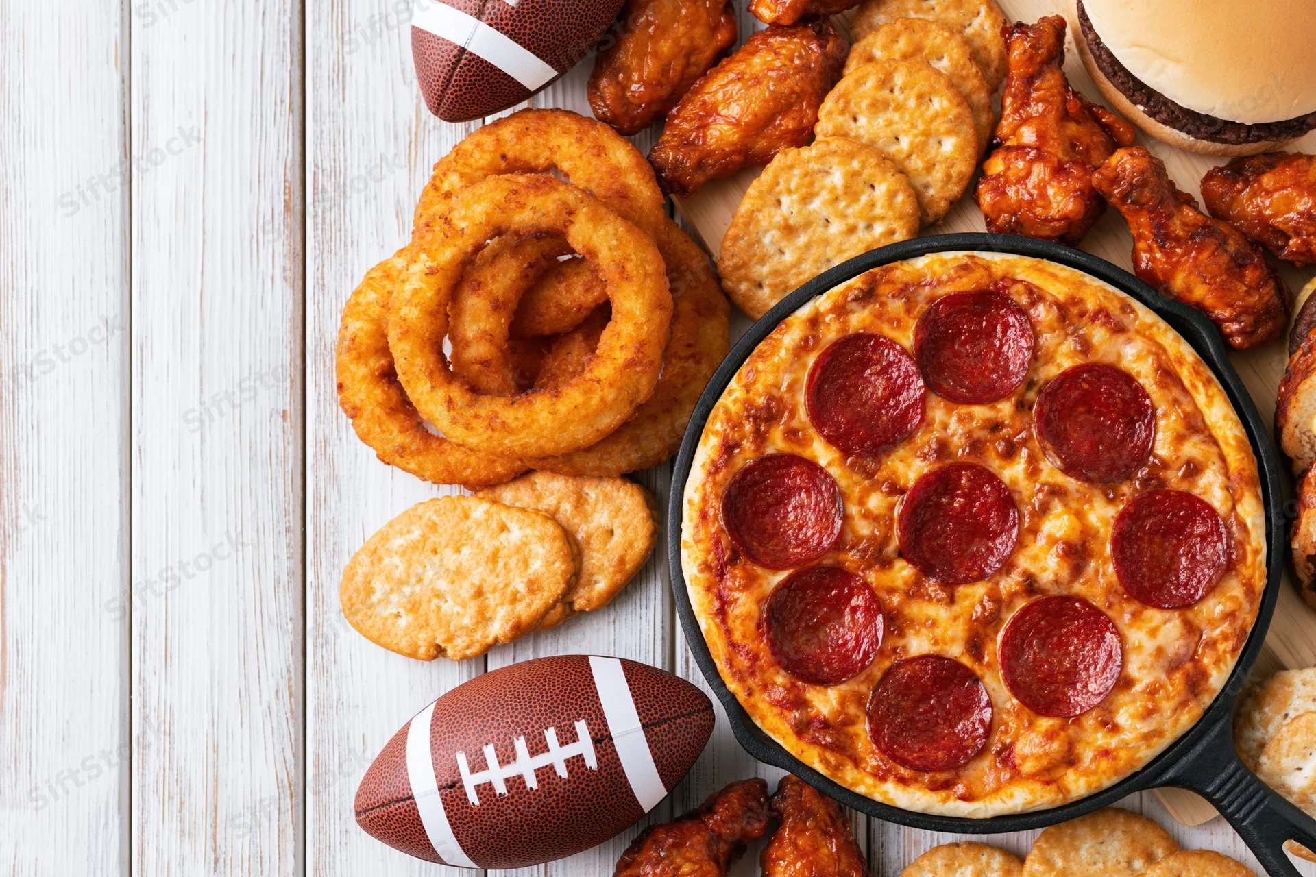 Game-day food spread with pizza, wings, onion rings and footballs on a white wooden table