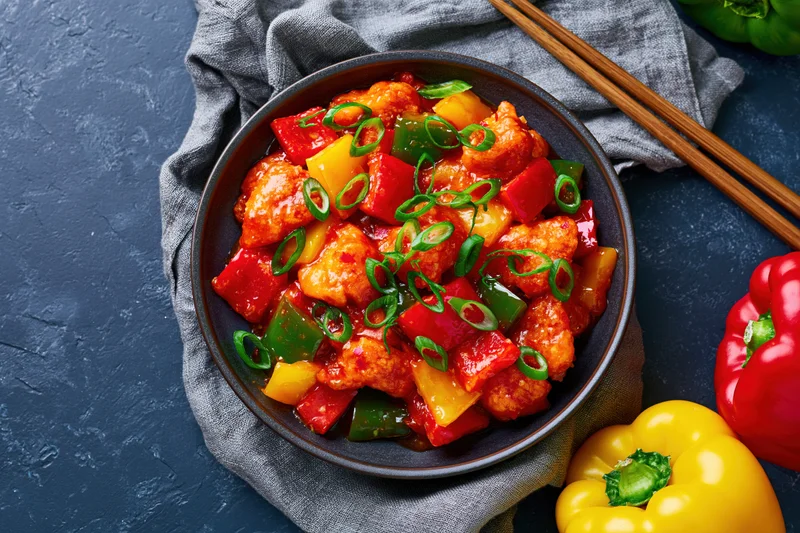 Sweet and sour chicken stir-fry in a bowl with bell peppers and pineapple, garnished with scallions, on a dark blue surface with chopsticks and napkin
