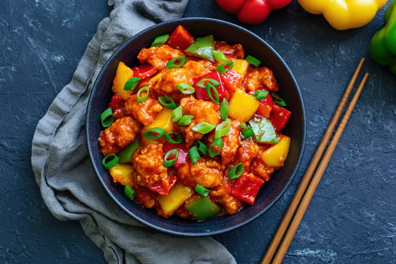Sweet and sour chicken stir-fry in a dark bowl with bell peppers and pineapple, served with chopsticks on a dark surface