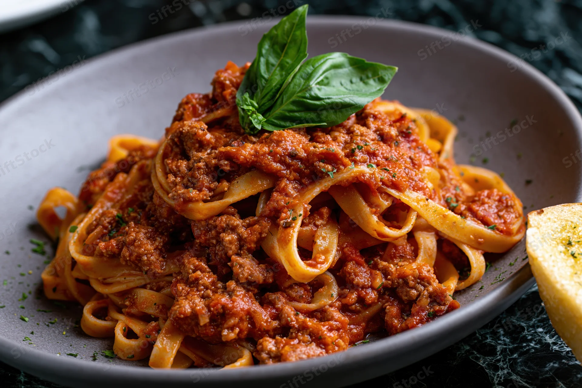 Tagliatelle bolognese served in a matte grey ceramic plate with basil garnish and garlic bread on the side