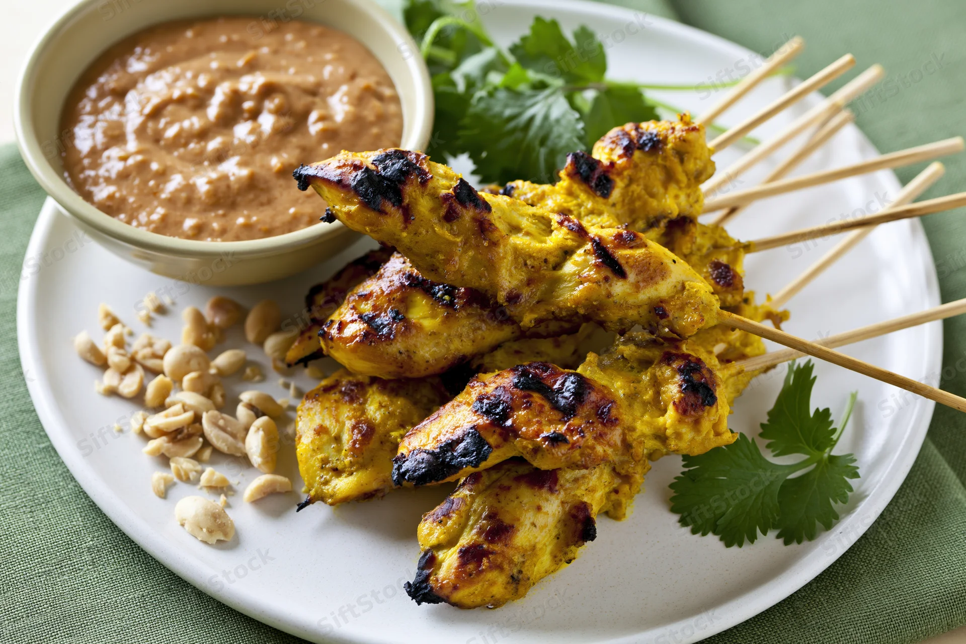 Grilled thai chicken satay skewers stacked on a white ceramic plate with peanut sauce and cilantro garnish