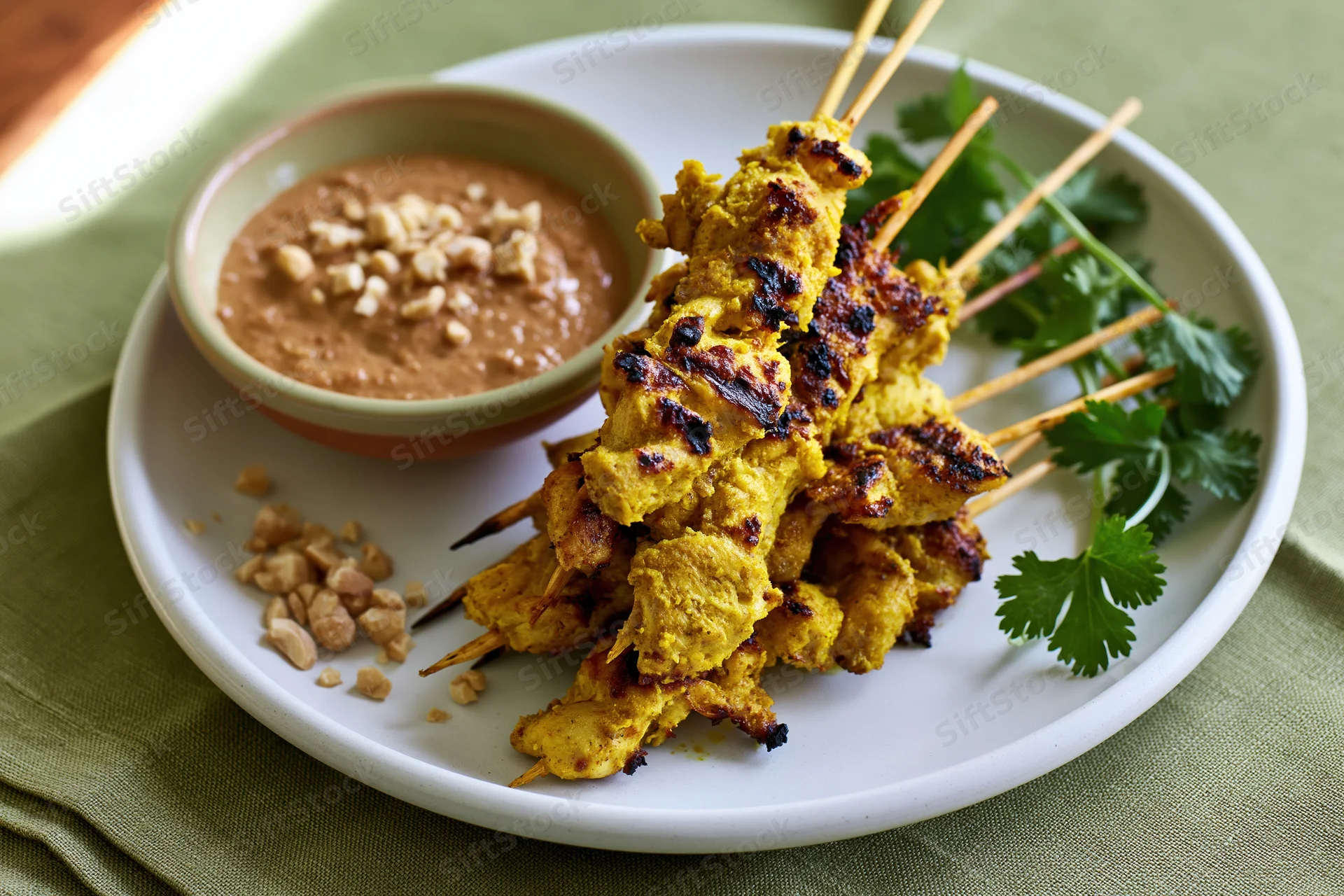 Thai chicken satay skewers stacked on a white ceramic plate with peanut sauce and cilantro garnish