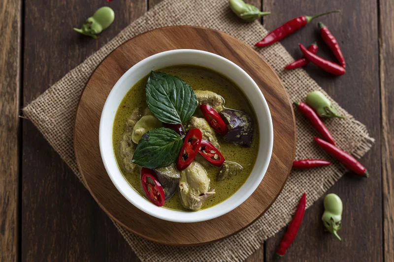 Thai green curry with chicken in a white ceramic bowl on a wooden tray with chili peppers and basil garnish