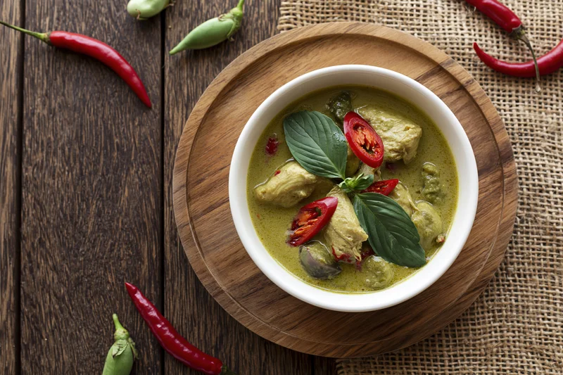 Thai green curry with chicken in a white ceramic bowl on a wooden tray with chili peppers and basil garnish