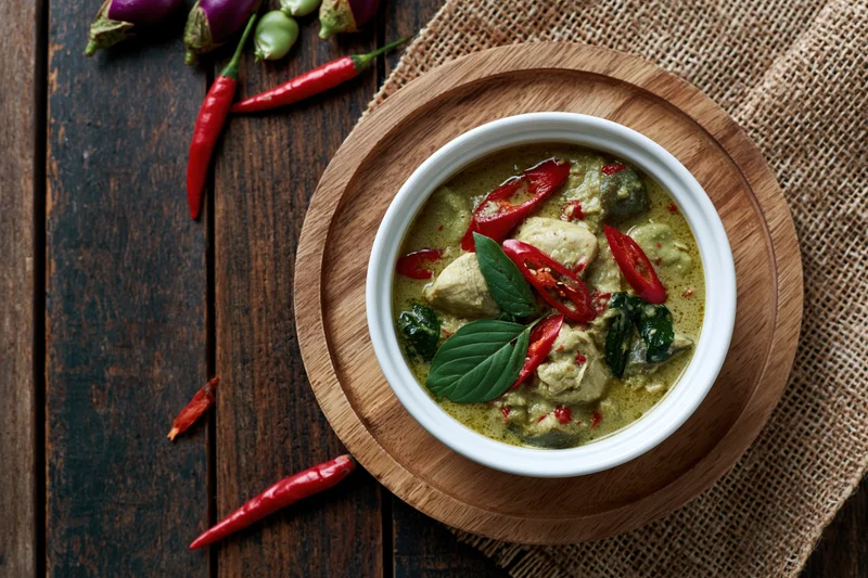 Thai green curry with chicken in a white ceramic bowl on a wooden tray with red chilies and basil leaves on a dark wood table