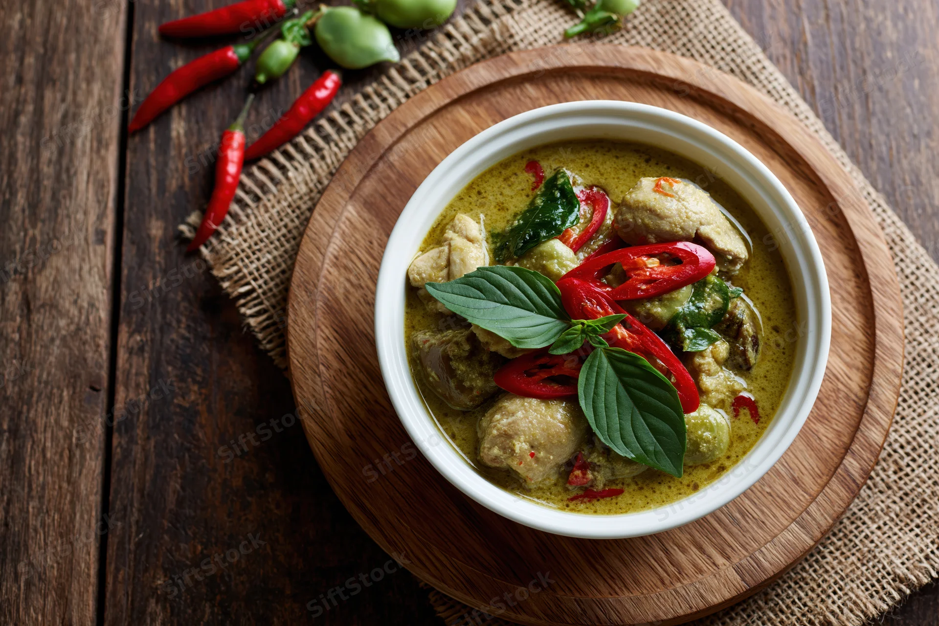 Thai green curry with chicken in a white ceramic bowl on a wooden tray with red chilies and basil leaves