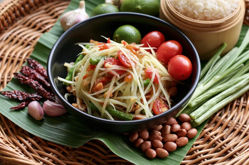 Thai green papaya salad in a black ceramic bowl surrounded by fresh ingredients on a banana leaf and woven basket