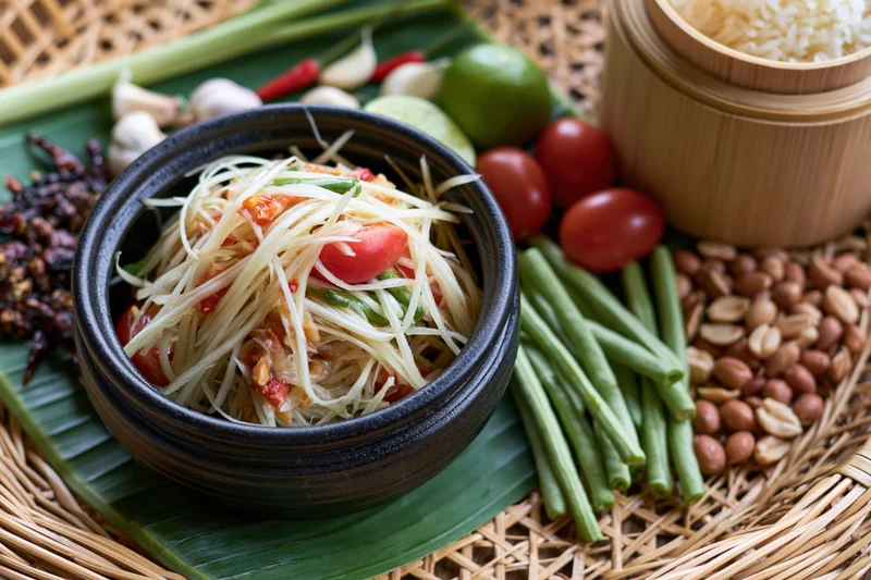 Thai green papaya salad in a black ceramic bowl surrounded by fresh ingredients on a banana leaf and woven tray