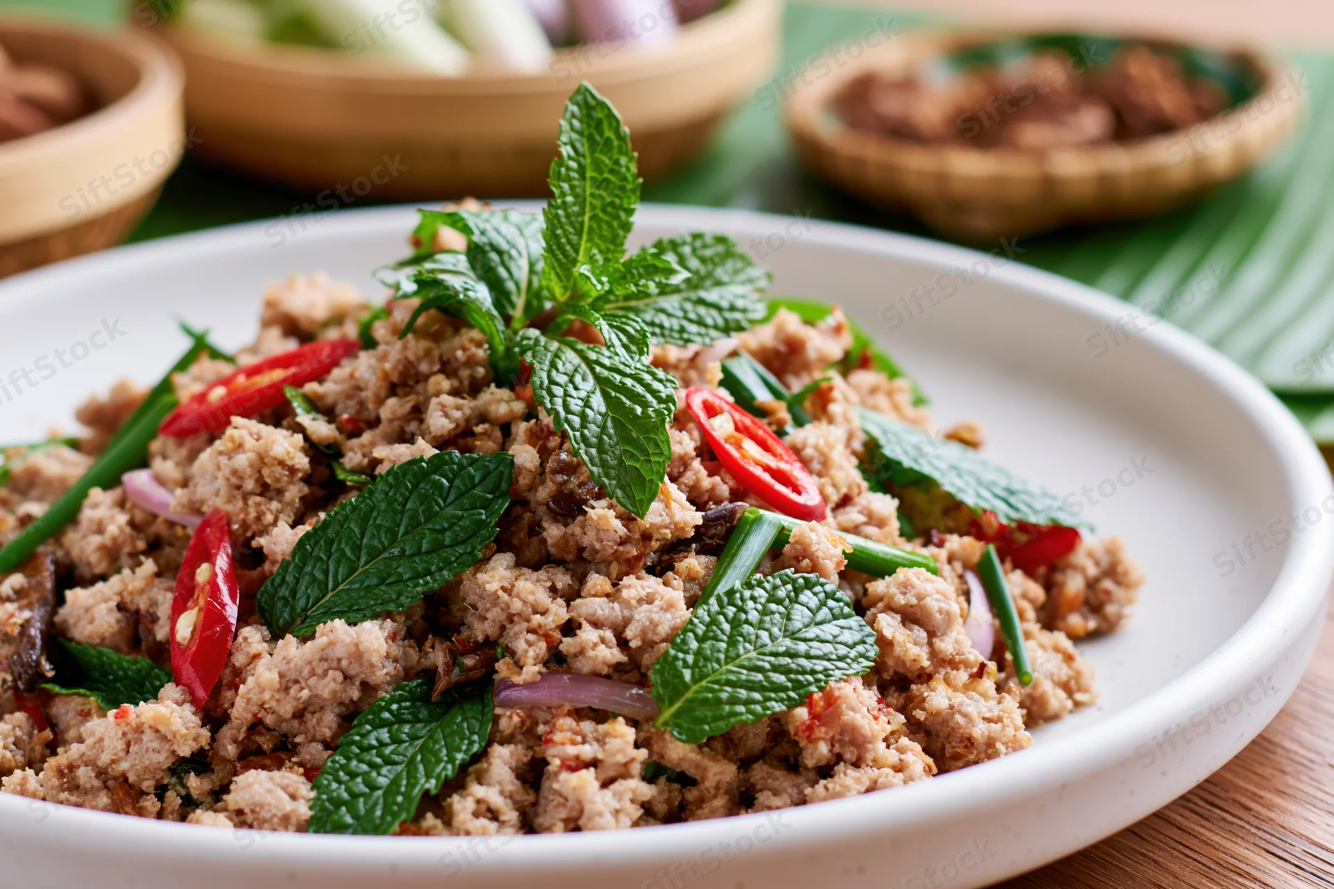 Thai larb moo minced pork salad garnished with mint and chili on a white plate with bamboo bowls in background