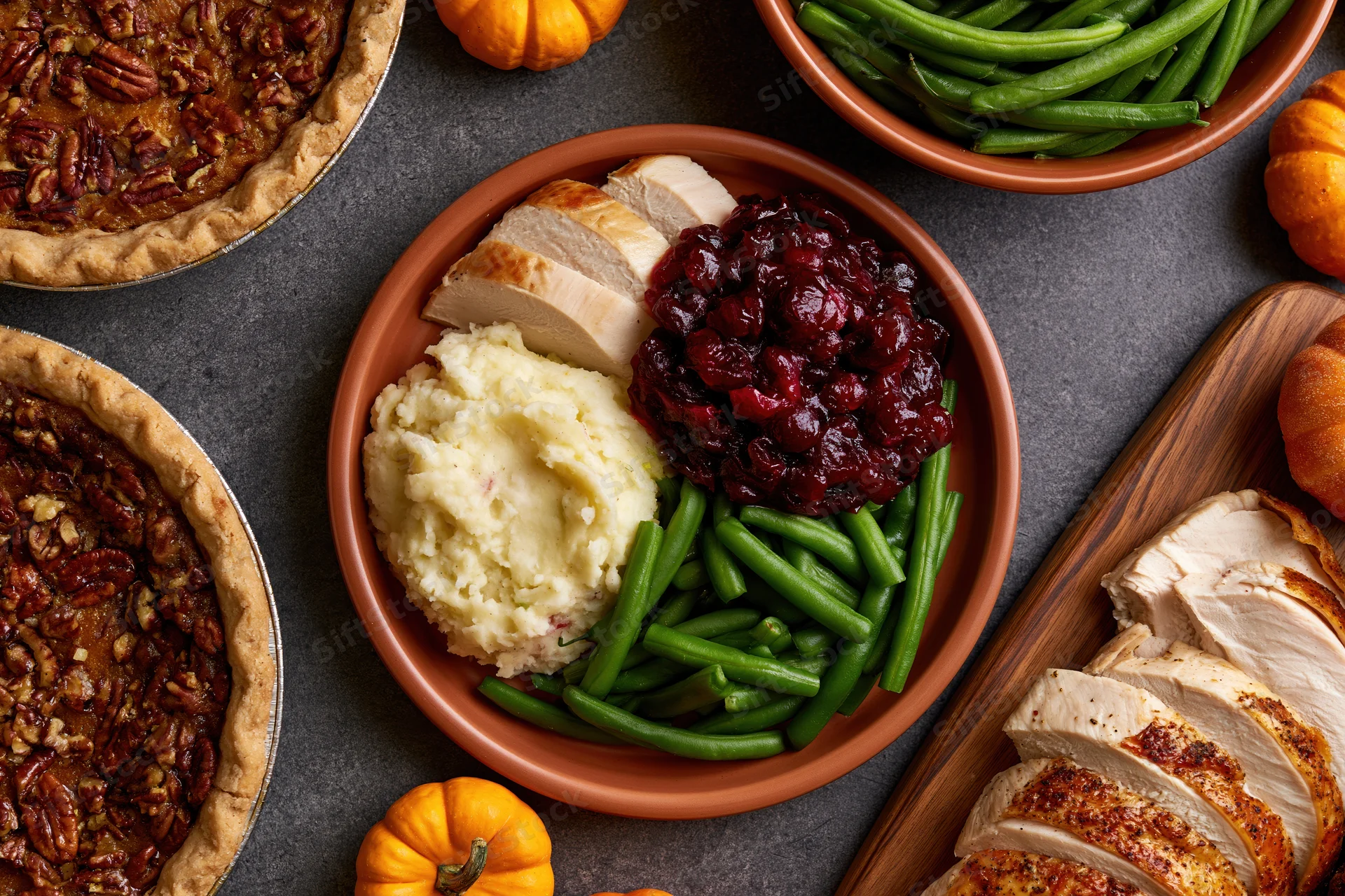 Thanksgiving dinner spread with turkey, cranberry sauce, mashed potatoes, green beans, and pecan pie on dark slate table