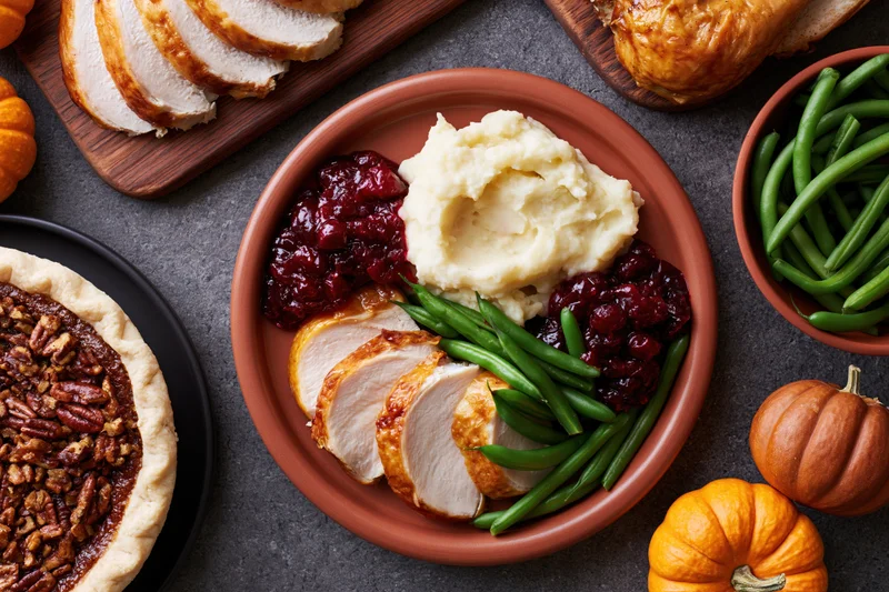 Thanksgiving dinner spread with turkey, mashed potatoes, cranberry sauce, green beans, and pecan pie on dark slate table with pumpkins and autumn decor
