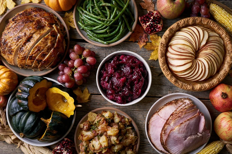 Thanksgiving feast spread with turkey, ham, cranberry sauce, green beans, apple pie, and autumn vegetables on a rustic wooden table
