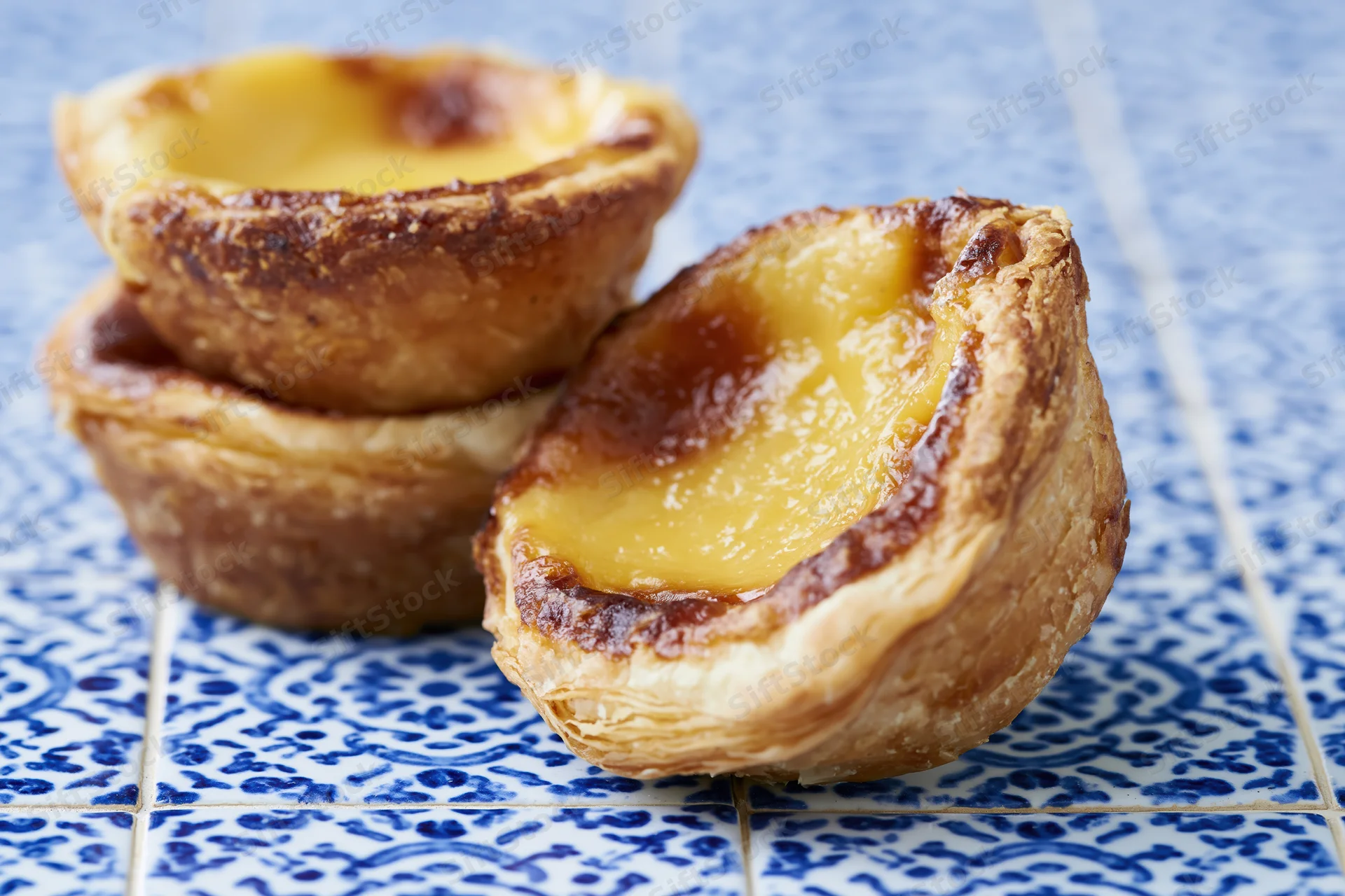 Three portuguese egg tarts with flaky crust and golden custard filling on blue patterned tiles