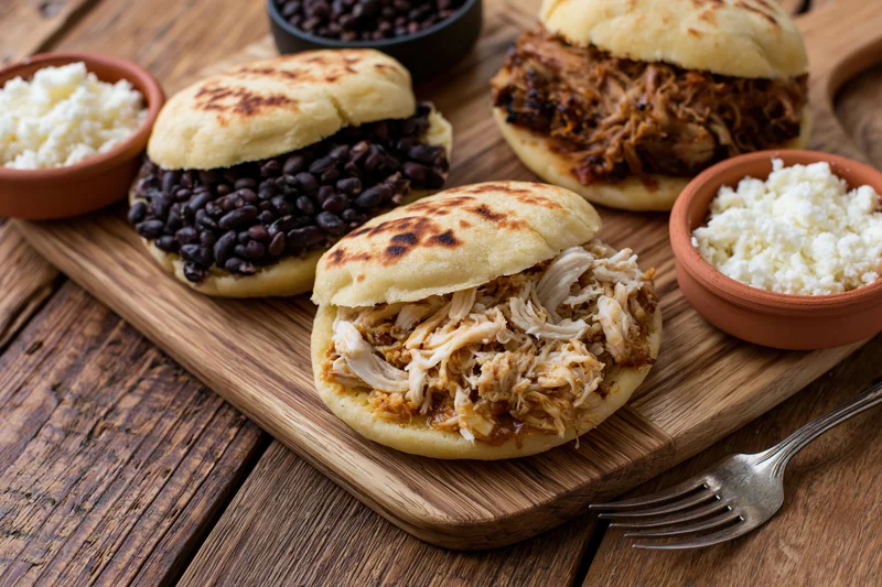 Three venezuelan arepas filled with shredded chicken and black beans served on a wooden board with cheese and bowls on the side