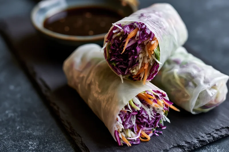 Three vietnamese fresh spring rolls with rice paper wrapping filled with purple cabbage carrots and lettuce on a dark slate plate beside a bowl of dipping sauce