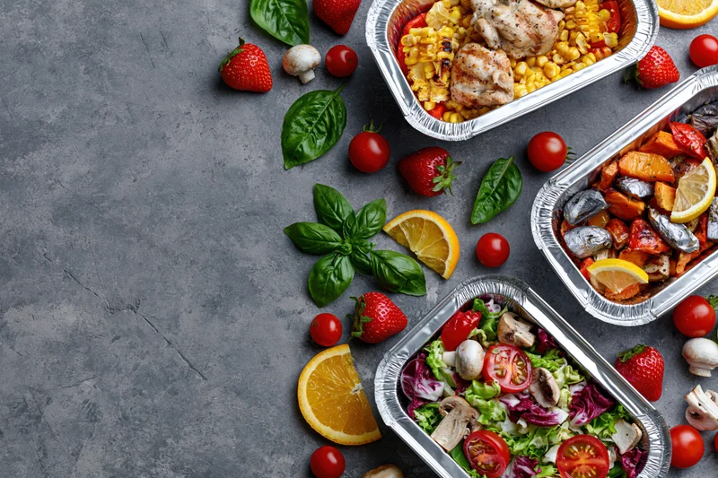 Three aluminum foil containers with grilled chicken, corn, salad and vegetables arranged on a gray surface with fresh fruit and herbs scattered around