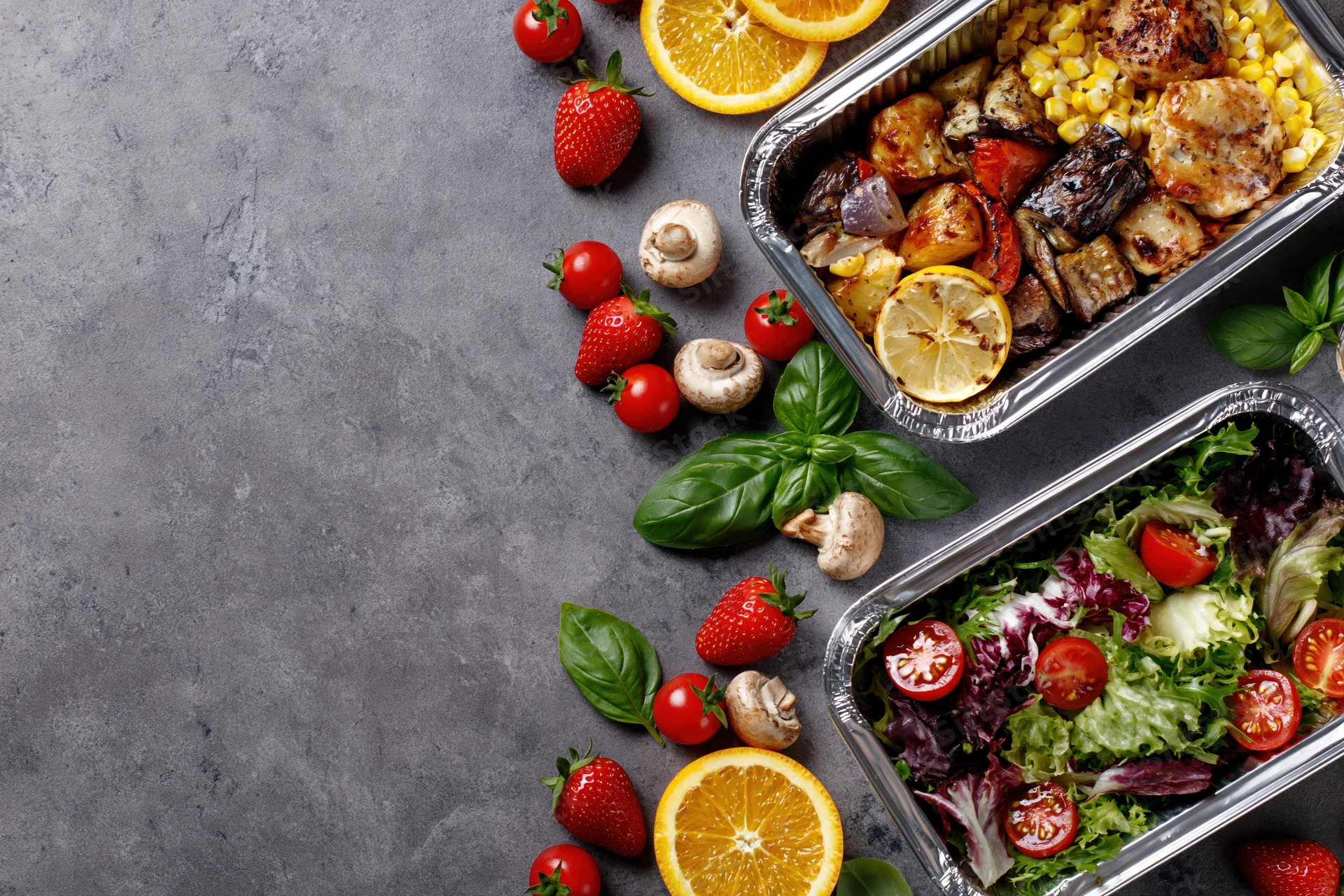 Two aluminum foil containers with grilled chicken and vegetables, and a salad, arranged on a gray surface with fresh fruit and herbs around