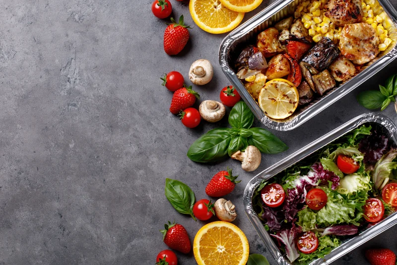 Two aluminum foil containers with grilled chicken and vegetables, and a salad, arranged on a gray surface with fresh fruit and herbs around