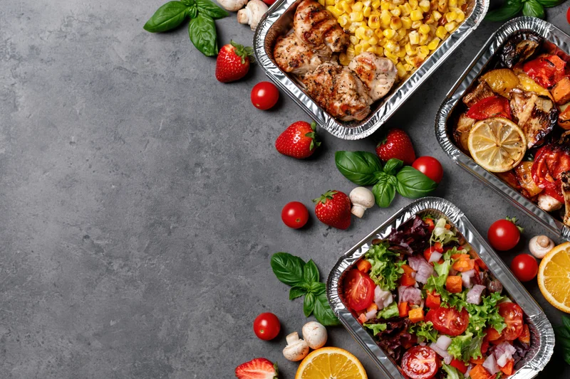 Three aluminum foil containers with grilled chicken, corn, salad and fresh vegetables arranged on a gray surface with scattered fruit and herbs