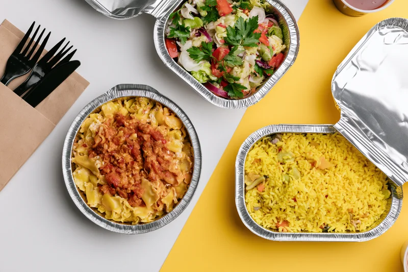 Three aluminum foil takeout containers with pasta bolognese, yellow rice, and salad on a white and yellow surface