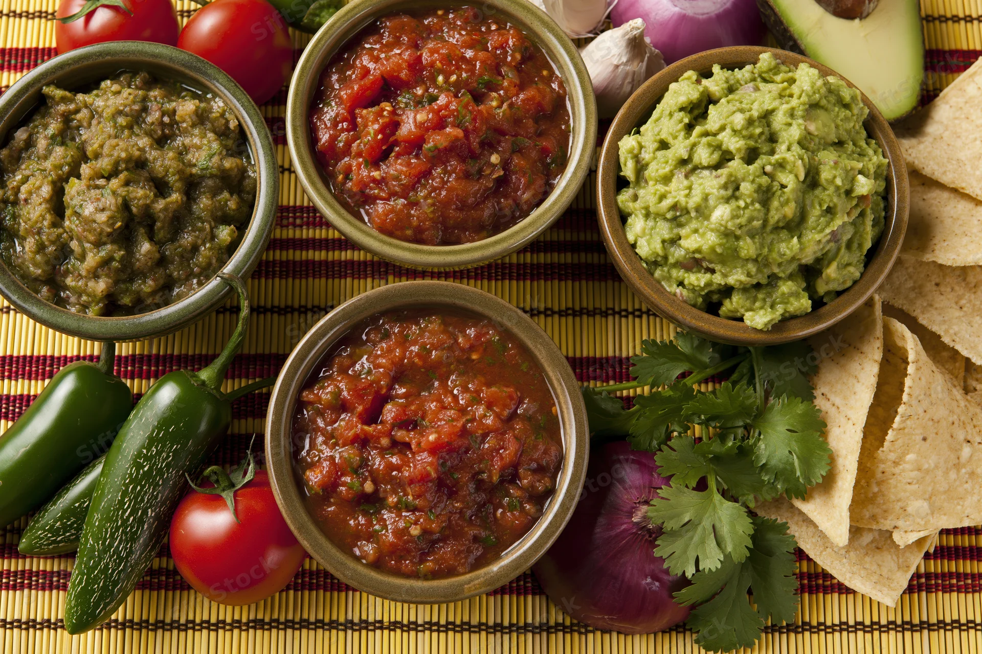 Three bowls of mexican salsa and guacamole with fresh vegetables and tortilla chips on a striped placemat