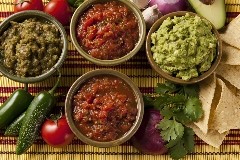 Three bowls of mexican salsa and guacamole with fresh vegetables and tortilla chips on a striped placemat