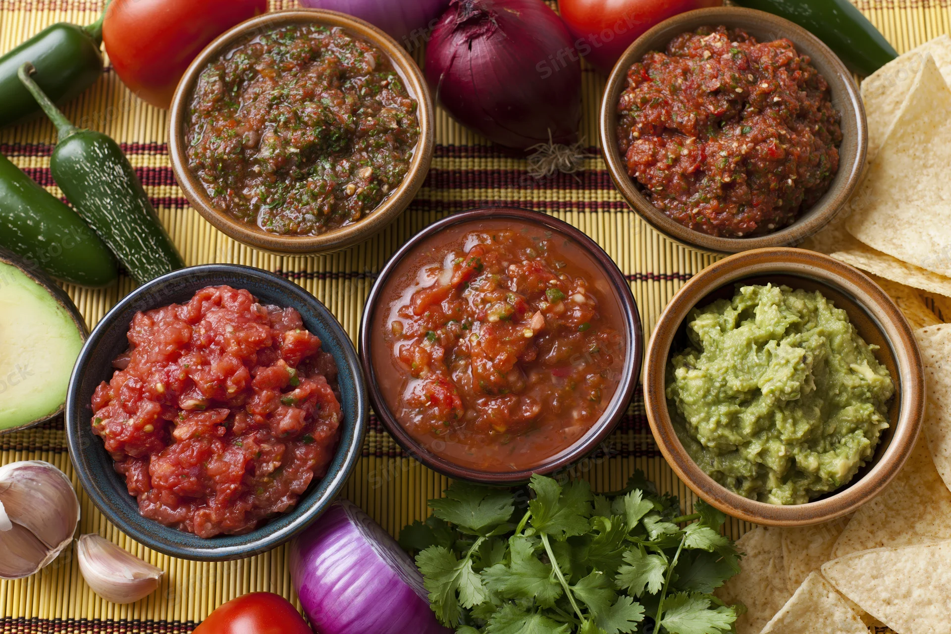 Several bowls of mexican salsas and guacamole arranged on a bamboo mat with fresh vegetables and tortilla chips around