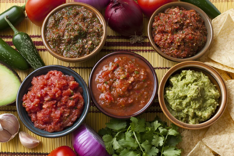 Several bowls of mexican salsas and guacamole arranged on a bamboo mat with fresh vegetables and tortilla chips around