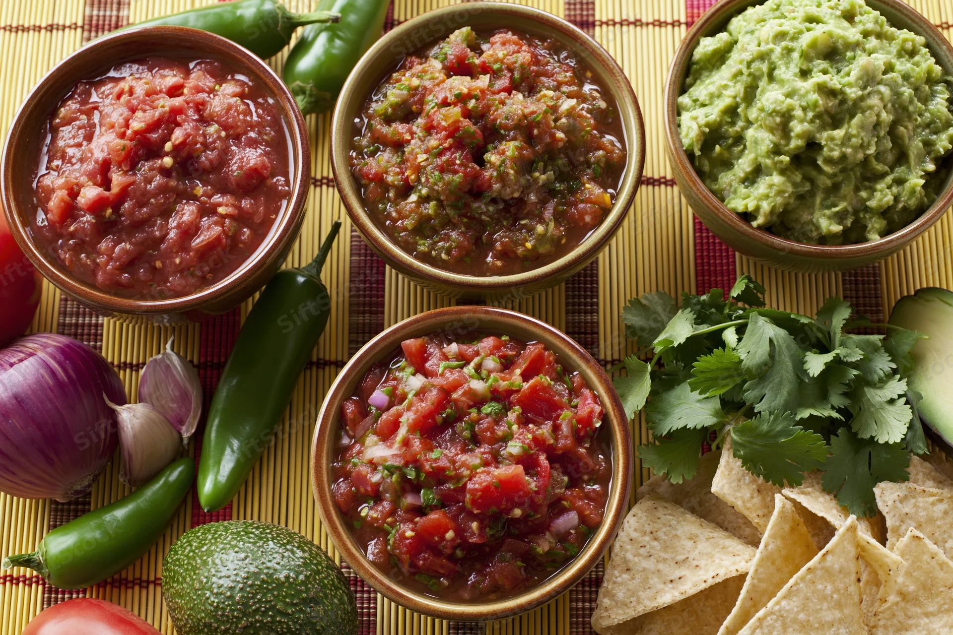 Three bowls of mexican salsa and guacamole with fresh ingredients arranged on a bamboo mat with tortilla chips