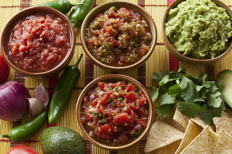 Three bowls of mexican salsa and guacamole with fresh ingredients arranged on a bamboo mat with tortilla chips